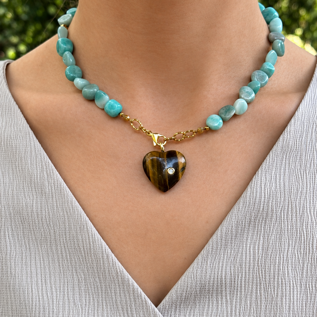 A close-up of a person wearing a beaded necklace with light blue-green turquoise beads, a gold chain, and a polished heart-shaped pendant with tiger-stripe patterns.